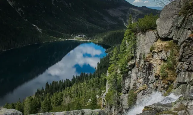 Gdzie jest Morskie Oko? Jezioro odbija chmury, otoczone lasem i górami. Wodospad spada po skalnej ścianie.
