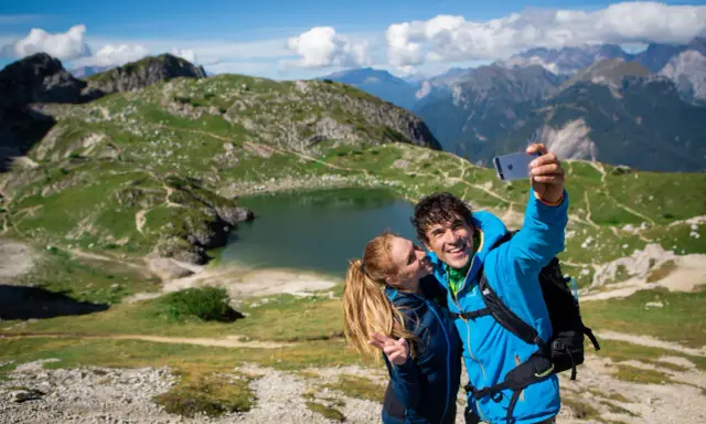 Coppia felice si scatta un selfie con il lago Coldai sullo sfondo, circondato da montagne verdi e un cielo azzurro con nuvole bianche.