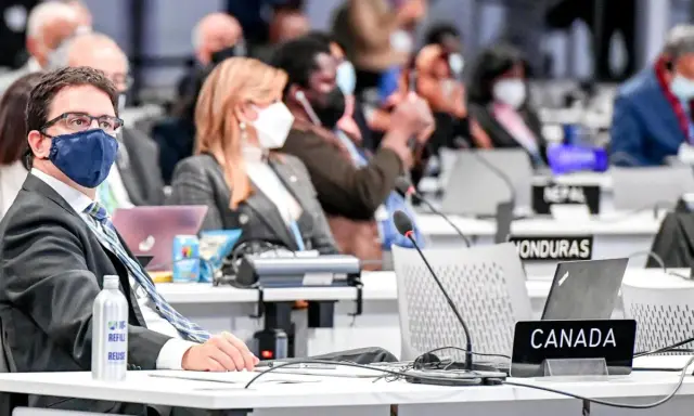 Teilnehmer der Klimakonferenz, darunter ein Mann mit Brille und Maske, sitzen an Tischen mit Laptops und Namensschildern wie "CANADA".