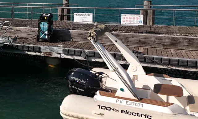 Un bateau électrique est prêt à être rechargé sur un ponton en bois. Une borne de recharge AQUA est visible à proximité.