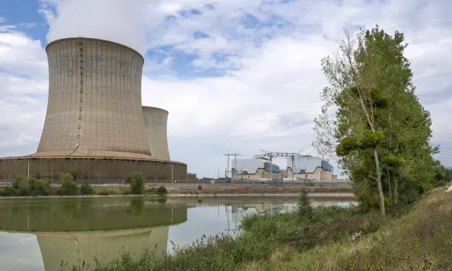 Kühltürme eines Atomkraftwerks in Frankreich, die Dampf abgeben. Ein ruhiger See spiegelt die Anlage wider, während am Ufer Bäume und Gras wachsen.