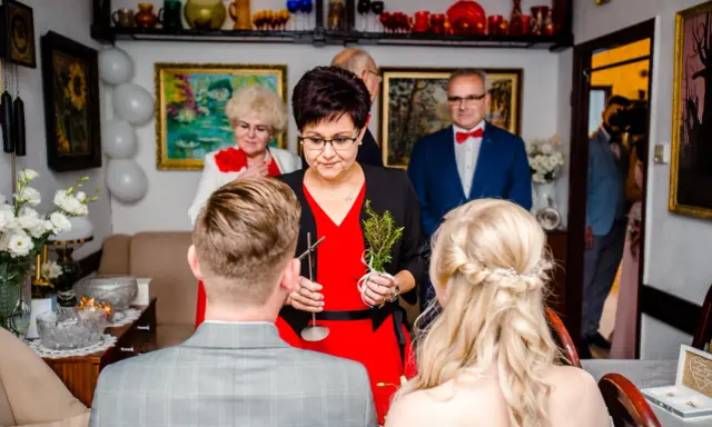 Rodzina udziela błogosławieństwa młodej parze. Kobieta trzyma gałązkę ziół, symbolizującą pomyślność.