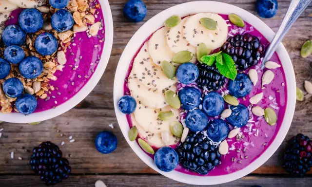 Zwei Schalen mit gesunden Bowls, gefüllt mit lila Smoothie, garniert mit Blaubeeren, Bananenscheiben, Brombeeren, Kürbiskernen und Granola.