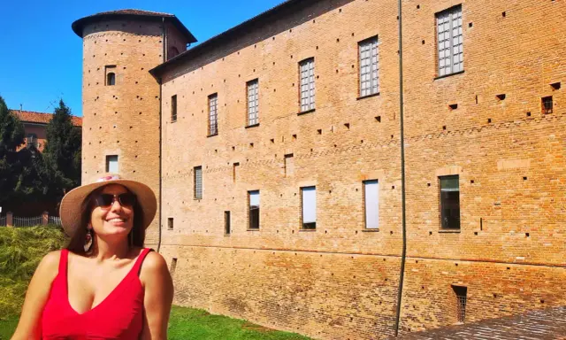 Donna sorridente con cappello di paglia davanti a un castello di mattoni rossi. Un'idea per cosa vedere a Piacenza in un giorno.