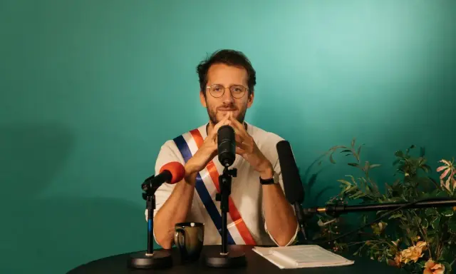 Un homme avec une écharpe tricolore, prêt à parler. Il est entouré de micros, peut-être pour un discours plein de langue de bois.