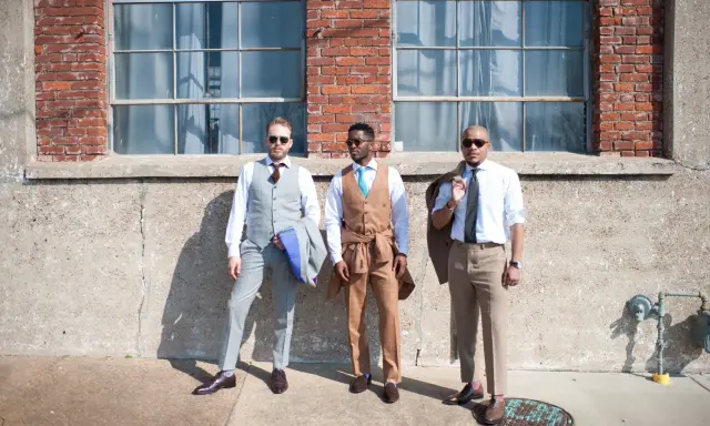 Three men in stylish suits embody cocktail chic attire. One wears a grey pinstripe suit, another a tan suit with a turquoise tie, and the third a light blue shirt with tan trousers.