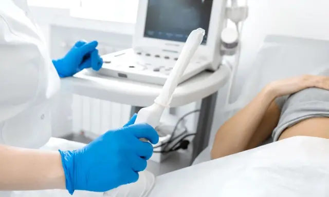 Medico con guanti blu tiene sonda per ecografia transvaginale. Paziente sdraiata sullo sfondo.