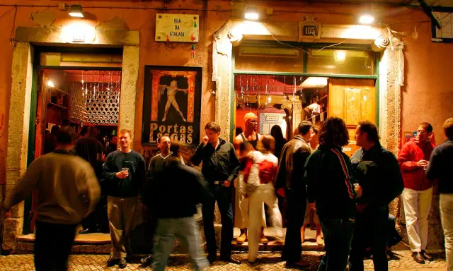 Menschen versammeln sich vor einem beleuchteten Lokal in Bairro Alto. Ein Mann hält ein Glas, andere unterhalten sich.