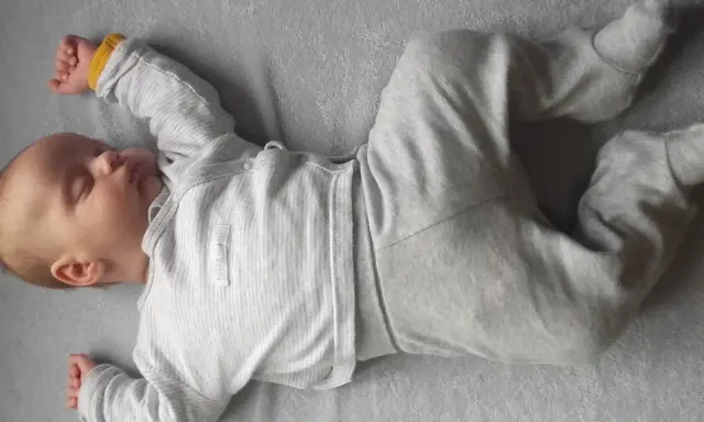Śpiący noworodek w szarym pajacyku. Jego rączki i nóżki są ułożone w lekkiej asymetrii, co jest naturalne dla niemowląt.