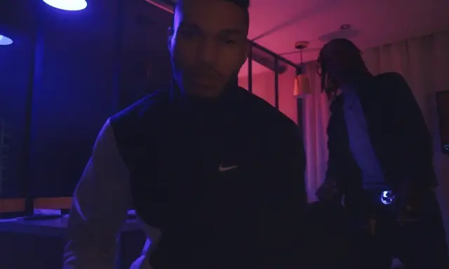 Dwóch mężczyzn w ciemnym pomieszczeniu, oświetlonym niebieskim i różowym światłem. Jeden z nich, 730 huncho, ma na sobie bluzę z logo Nike.