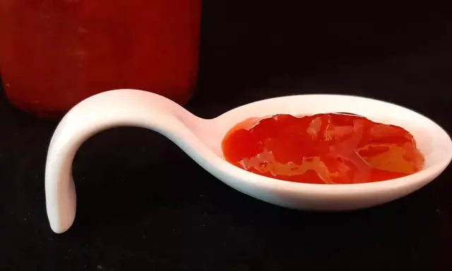 Une cuillère blanche présente une portion de confiture fraise rhubarbe, d'un rouge vif et appétissant. Un pot de confiture est visible en arrière-plan.