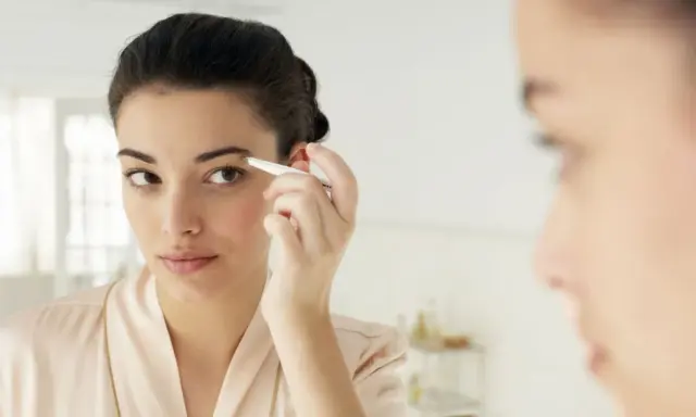 Une jeune femme ajuste ses sourcils broussailleux avec une pince à épiler devant un miroir, dans une tenue de chambre satinée.