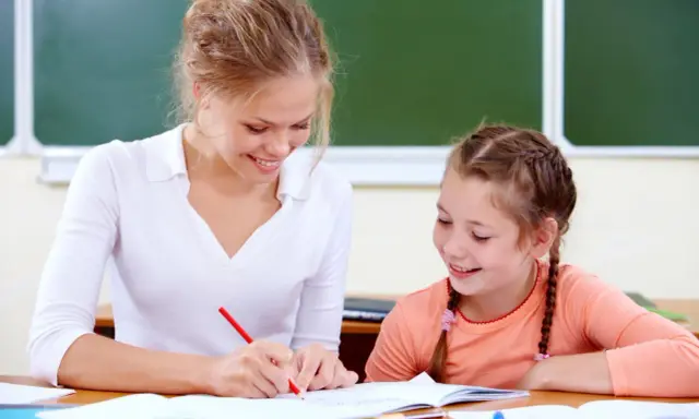Uśmiechnięta nauczycielka wspomagająca z kwalifikacjami pomaga uczennicy w lekcjach przy biurku.