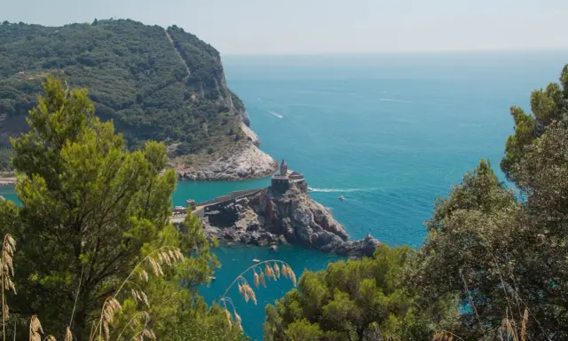 Vista panoramica del sentiero Riomaggiore Portovenere, con la chiesa di San Pietro su uno sperone roccioso e il mare azzurro.