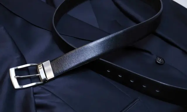 A black leather belt with a silver buckle rests on a dark navy suit jacket. Do you wear a belt with a tuxedo? This image shows a classic accessory.