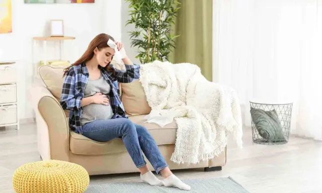 Donna incinta seduta sul divano con un fazzoletto sulla fronte, che soffre di raffreddore in gravidanza.