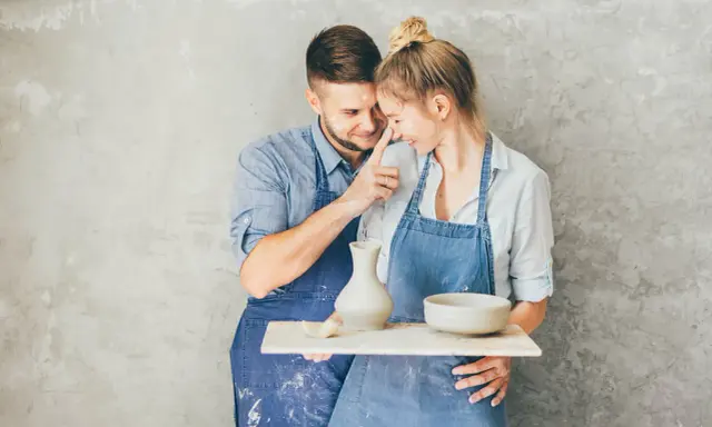 Para świętuje 9 rocznicę ślubu, tworząc ceramikę. Mężczyzna delikatnie dotyka nosa kobiety, oboje ubrani w fartuchy, ubrudzeni gliną.