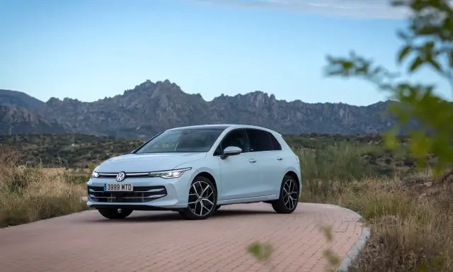 Un coche compacto Volkswagen Golf blanco circula por una carretera de adoquines con montañas rocosas al fondo.