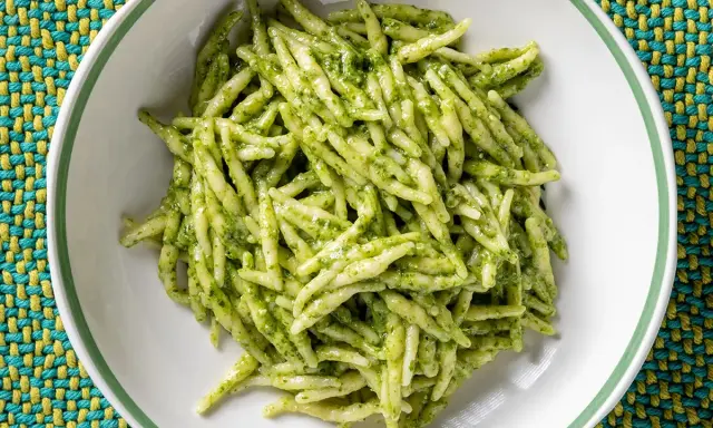 Un piatto di trofie al pesto, pasta fresca condita con un sugo verde brillante, servita in una ciotola bianca su una tovaglietta colorata.