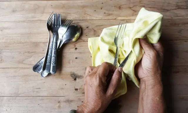 Mains polissant des couverts en argent avec un chiffon jaune. Un tas de fourchettes et cuillères est à gauche. Apprenez comment nettoyer l'argenterie.