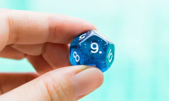 Palec i kciuk trzymają niebieską, dwudziestościenną kostkę z błyszczącymi drobinkami. Widoczna jest cyfra 9, symbolizująca numerologiczną 9.