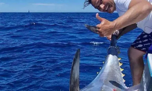 Uomo felice mostra il suo **biggest tuna** appena pescato in mare aperto, con una barca e una vela all'orizzonte.