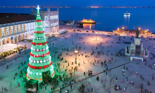Lisbona illuminata per il weekend dell'Immacolata: un imponente albero di Natale brilla in una piazza affollata, con il fiume e le luci della città sullo sfondo.