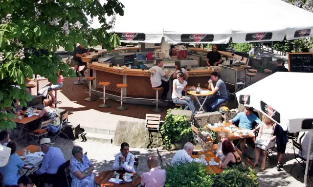 Gäste sitzen an Tischen und an der Theke eines Biergartens unter Sonnenschirmen.