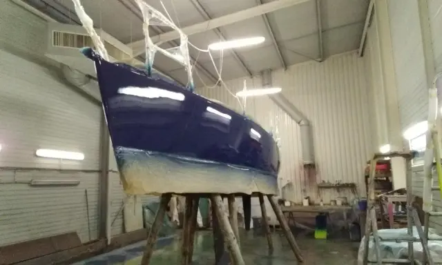 Un bateau bleu profond est en cours de stratification dans un atelier. La coque est soutenue par des tréteaux en bois.
