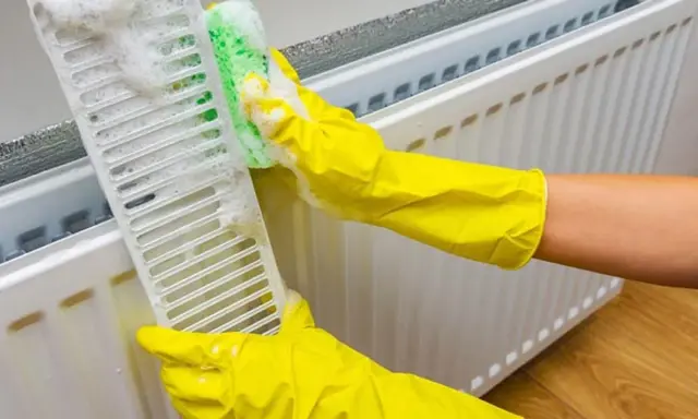 Manos con guantes amarillos limpian la rejilla de un radiador con una esponja y espuma. Guía para saber como limpiar radiador.