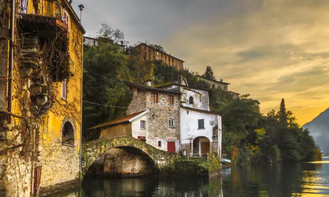 Malerische Häuser am Wasser in Nesso am Comer See, mit einer alten Steinbrücke und goldenem Abendlicht.