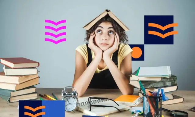 Zmęczona studentka z książką na głowie, otoczona stosami książek i materiałami do nauki. Tak wyglądają studia niestacjonarne.