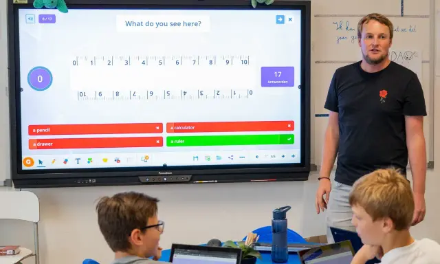 Un insegnante guida una lezione interattiva, simile a Kahoot, con domande su uno schermo touch. I bambini partecipano con entusiasmo.