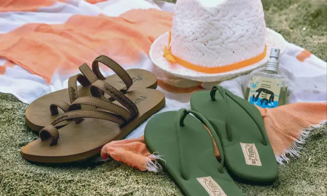 Brown sandals vs. green flip flops on a beach towel, with a hat and body mist. A perfect beach day awaits!