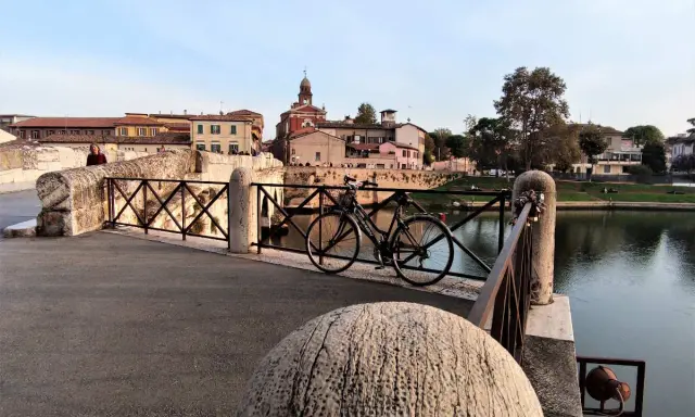 Ponte sul fiume a Borgo San Giuliano, con una bicicletta appoggiata alla ringhiera e palazzi sullo sfondo.