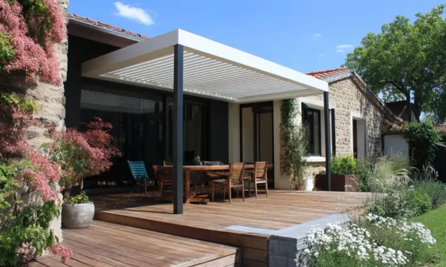Construction d'une pergola moderne sur une terrasse en bois, avec une table et des chaises, devant une maison en pierre.