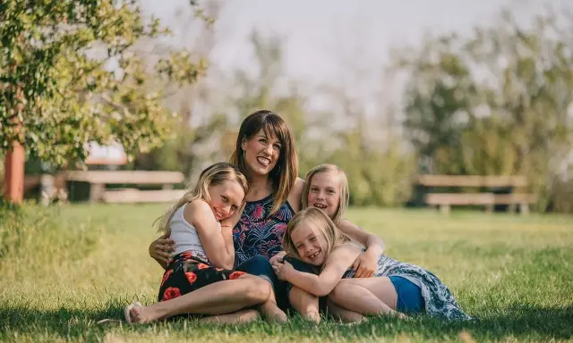 Mama z córkami na trawie, uśmiechnięte i przytulone. Widać, że czują motyle w brzuchu od tej miłości.