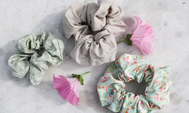 Tre virka scrunchies i olika mönster och färger, omgivna av rosa blommor på en marmorerad yta.