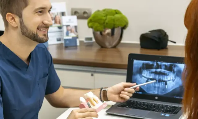 Dentysta omawia z pacjentką plan leczenia, pokazując na ekranie zdjęcie rentgenowskie szczęki i model zębów, wyjaśniając znaczenie czyszczenia dziąseł.