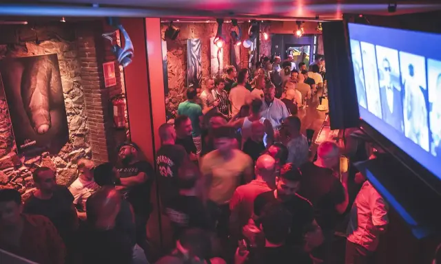 Un locale affollato a Barcellona by night, con uomini che socializzano e bevono. L'atmosfera è vibrante, illuminata da luci rosse e blu.