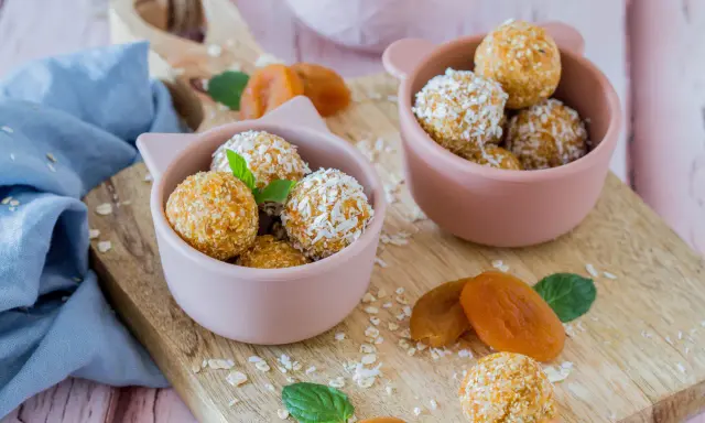 Zwei rosa Schalen gefüllt mit leckeren Energy Balls, bestreut mit Kokosraspeln und Haferflocken. Getrocknete Aprikosen und Minzblätter liegen daneben.