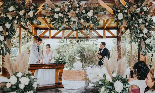 Novios celebran aniversario en altar decorado con flores y pampas. Un oficiante habla mientras la pareja sonríe.