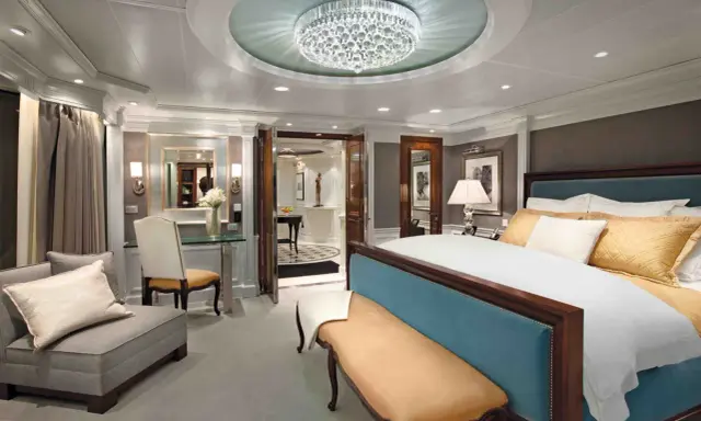 Lussuosa suite crociera con letto king-size, divanetto, lampadario di cristallo e vista su un corridoio elegante.