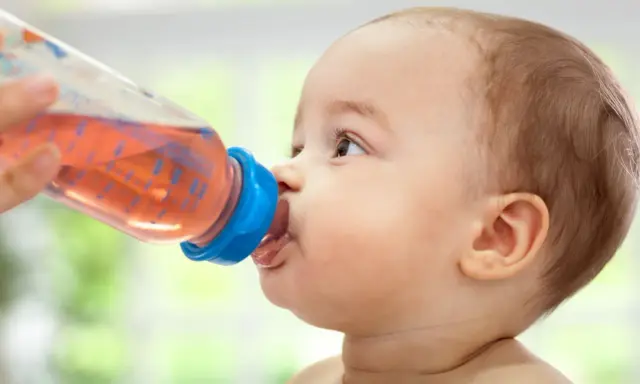 Kleines Baby trinkt aus einer Flasche mit rosafarbenem Tee. Ab wann Tee Baby? Diese Frage stellen sich viele Eltern.