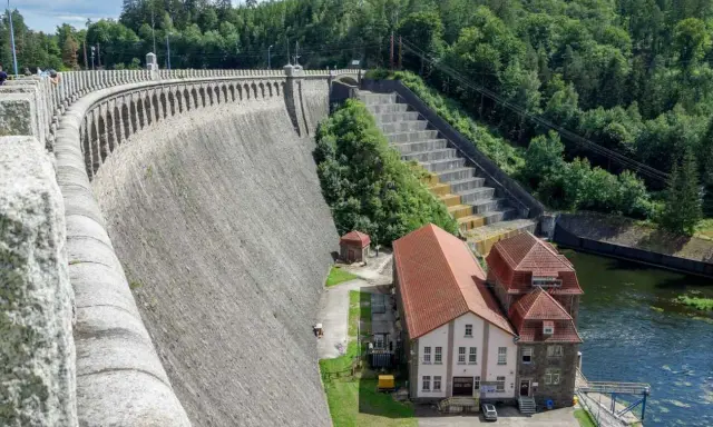 Potężna, kamienna zapora, największa zapora w Polsce, wznosi się nad rzeką. Obok niej budynki elektrowni i zielone wzgórza.