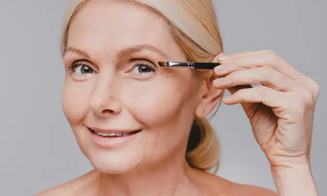 Une femme blonde applique du maquillage sur son œil, son sourcil blanc est visible.