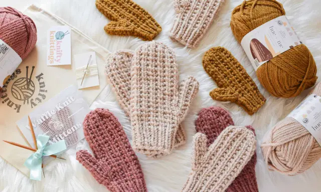 Mitones tejidos a mano con patrón de dos agujas. Varios pares en tonos rosa, beige y mostaza, junto a ovillos de lana y agujas.