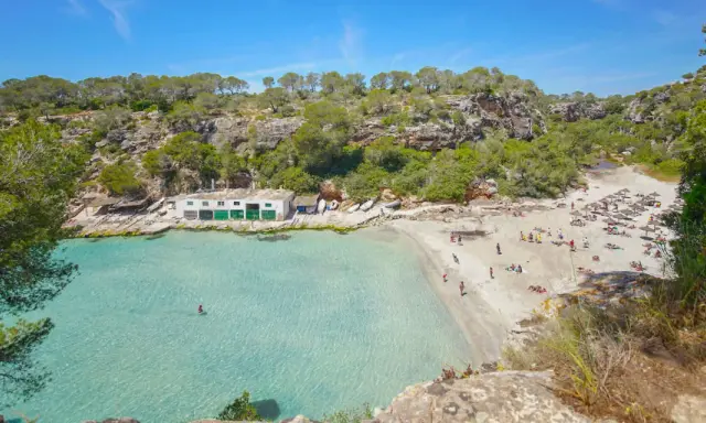 Türkisfarbenes Wasser und ein sonniger Sandstrand in Cala Pi, umgeben von grünen Hügeln. Menschen entspannen sich unter Sonnenschirmen.