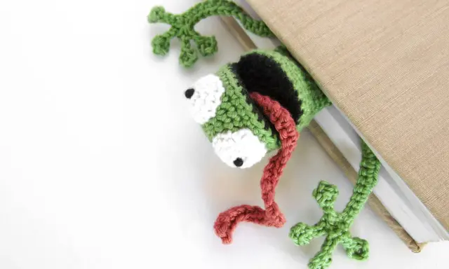 Marcador de libro con ranas amigurumi, verde y negro, con lengua roja, asomando de un libro beige.