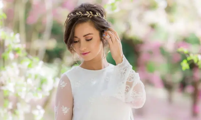 Delikatna tiara na głowę zdobi brunetkę w białej sukni z koronkowymi rękawami, otoczoną kwitnącymi drzewami.